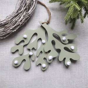 Op de afbeelding: Een saliegroene houten kerstversiering in de vorm van een maretak, versierd met kleine witte pompons. De versiering hangt aan een natuurlijke touwtje. Een rustieke krans en een takje groenblijvend zijn op de achtergrond.