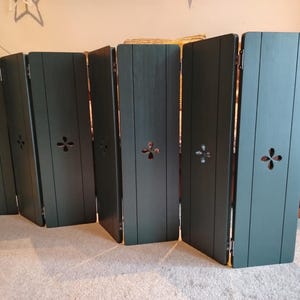 Op de afbeelding: Een donkergroen kamerscherm met zes panelen. Elk paneel heeft een decoratieve uitsparing en verticale houten latten. Het scherm is scharnierend en staat op een lichtgekleurd tapijt.