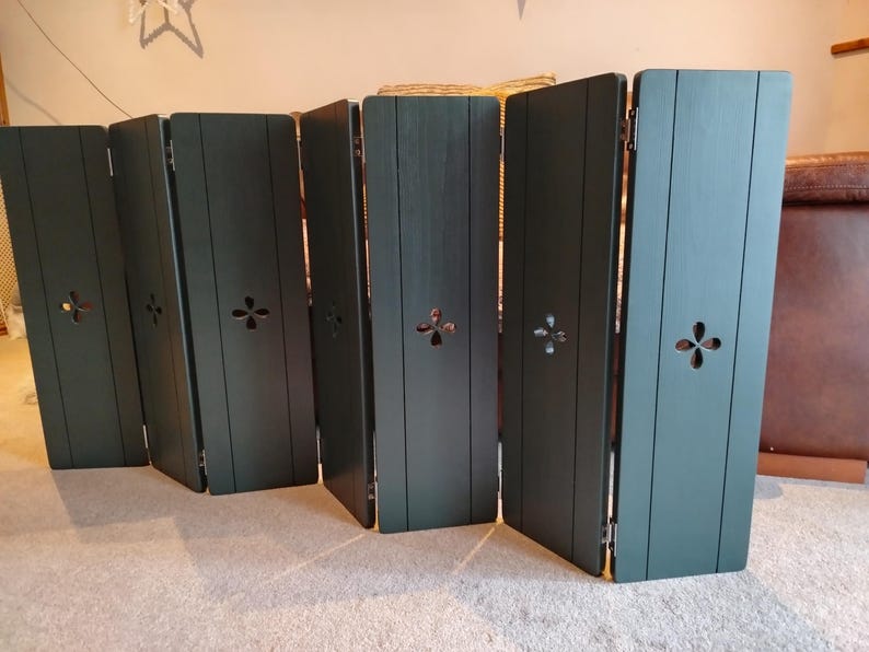 Op de afbeelding: Een donkergroen kamerscherm met zes panelen. Elk paneel heeft een decoratieve uitsparing en verticale houten latten. Het scherm is scharnierend en staat op een lichtgekleurd tapijt.