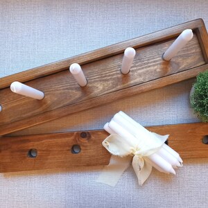 Op de afbeelding: Twee houten kaarsenstandaards, een met vijf gleuven voor kaarsen en de andere met drie gleuven. De standaards zijn in een donkerbruine kleur gebeitst. Een bundel witte kaarsen is vastgebonden met een wit lint.