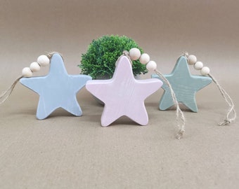 Può includere: Tre stelle ornamentali in legno con finitura rustica. Le stelle sono dipinte in azzurro, rosa e verde. Ogni stella ha una corda di perline di legno attaccata.