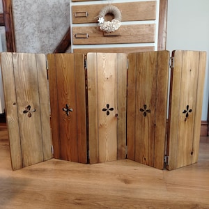 Op de afbeelding: Een houten kamerscherm met vijf panelen en een natuurlijke afwerking. Elk paneel heeft een decoratieve uitsparing en is verbonden door scharnieren. De houtnerf is zichtbaar. Het kamerscherm staat op een houten vloer.
