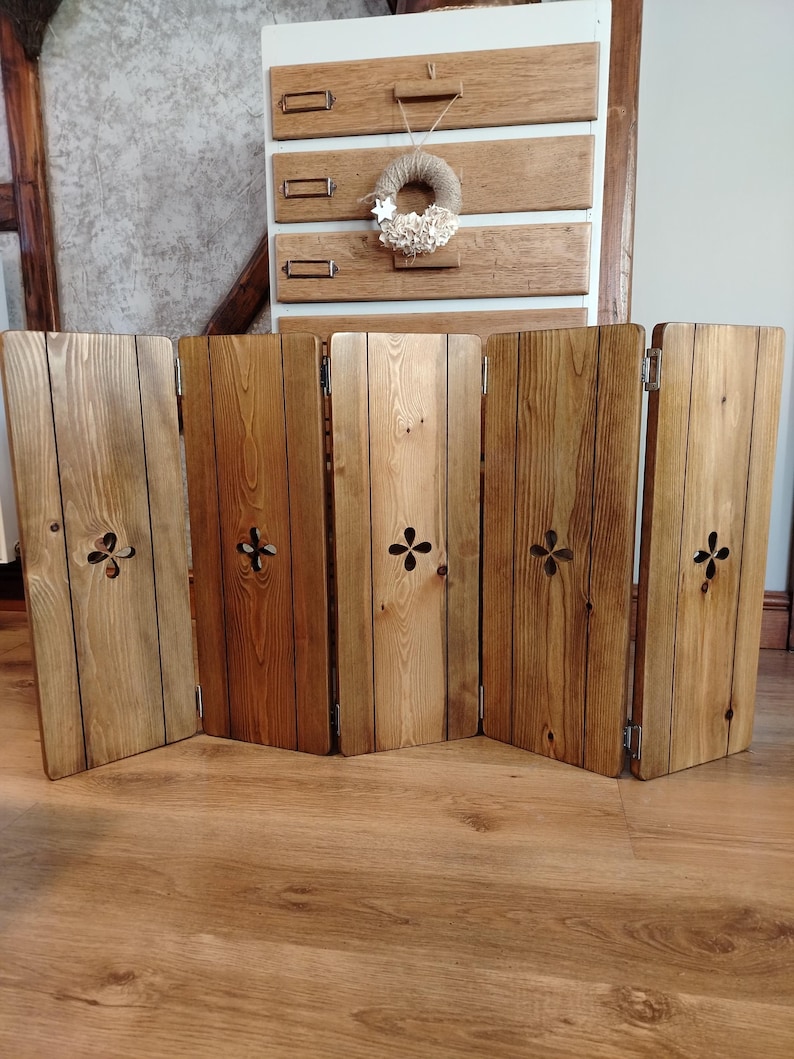 Op de afbeelding: Een houten kamerscherm met vijf panelen en een natuurlijke afwerking. Elk paneel heeft een decoratieve uitsparing en is verbonden door scharnieren. De houtnerf is zichtbaar. Het kamerscherm staat op een houten vloer.