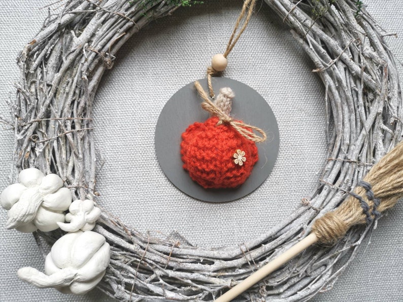 Pu&ograve; includere: Un ornamento a forma di zucca di maglia rossa con un fiocco di spago marrone e un piccolo accento di fiore di legno, appeso a una ghirlanda grigia con una base di ramoscelli marroni.