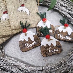 May include: Four wooden Christmas ornaments shaped like Christmas pudding with white icing and brown sauce. Each ornament has a different name written on it: Brian, Jason, Christina, and Amy.