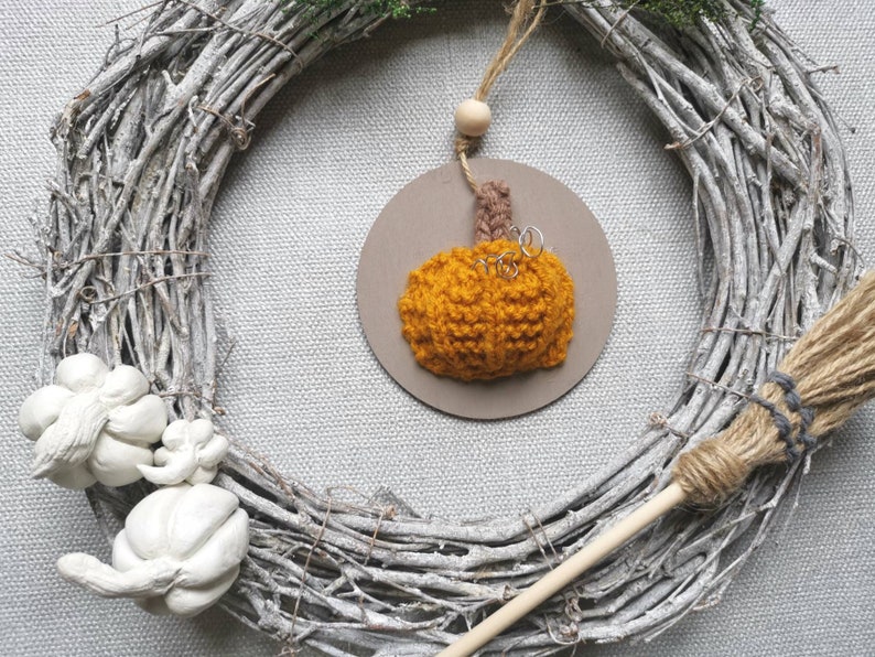 Pu&ograve; includere: Un ornamento a forma di zucca arancione lavorato a uncinetto appeso a una ghirlanda di rami bianchi con una scopa marrone e beige in primo piano.