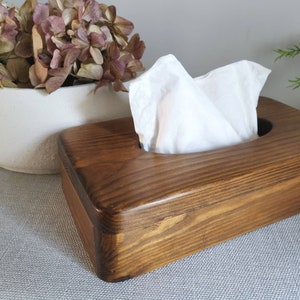 May include: A wooden tissue box holder with a dark brown stain. The box has a rectangular shape with rounded edges and a cutout for the tissues. The box is sitting on a light gray surface.