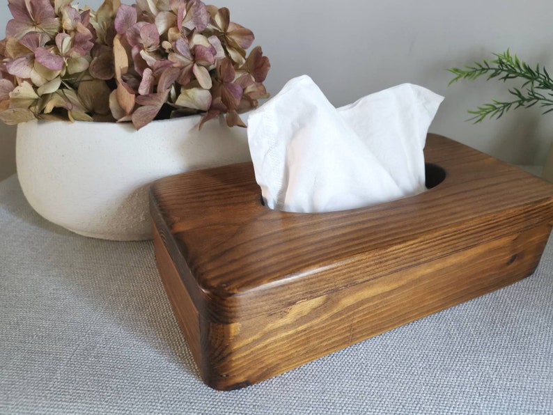 May include: A wooden tissue box holder with a dark brown stain. The box has a rectangular shape with rounded edges and a cutout for the tissues. The box is sitting on a light gray surface.
