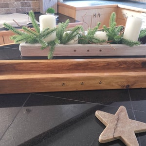 Op de afbeelding: Een houten plantenbak met een natuurlijke houten afwerking. De plantenbak heeft een rechthoekige vorm en is versierd met kleine stervormige details. De plantenbak staat op een zwart oppervlak.