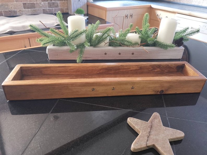 Op de afbeelding: Een houten plantenbak met een natuurlijke houten afwerking. De plantenbak heeft een rechthoekige vorm en is versierd met kleine stervormige details. De plantenbak staat op een zwart oppervlak.