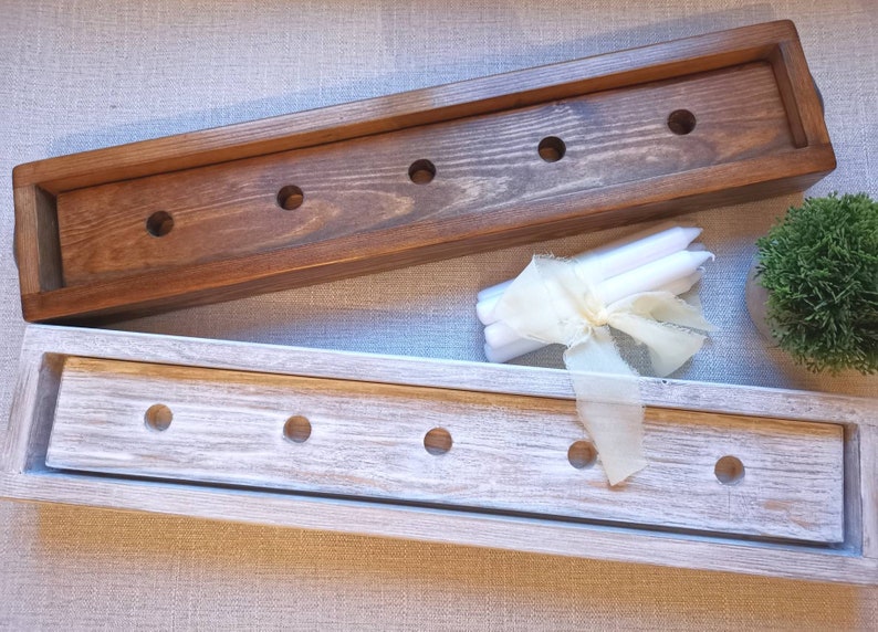 Op de afbeelding: Twee houten kaarsenhouders, een in bruin gebeitst en een in wit geverfd, met gaten voor kaarsen. De houders zijn leeg en er liggen witte kaarsen en een wit lint op de tafel.