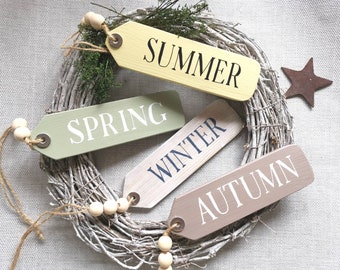 Può includere: Quattro etichette di legno con spago e perline bianche appese a una ghirlanda di rami bianchi. Le etichette sono dipinte in diversi colori e hanno le parole "Primavera", "Estate", "Autunno" e "Inverno" scritte su di esse.