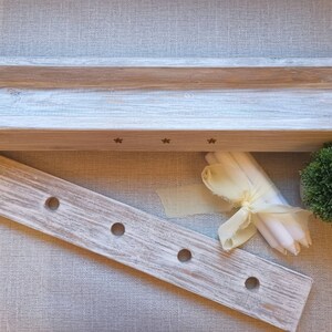 Op de afbeelding: Een witgekalkte houten doos met een rechthoekige opening en drie sterren die in de voorkant zijn gegraveerd. De doos staat op een lichtbruin stofoppervlak. Naast de doos staat een houten dienblad met vijf gaten. Een bundel witte kaarsen en een klein groen struikje liggen ook op het oppervlak.