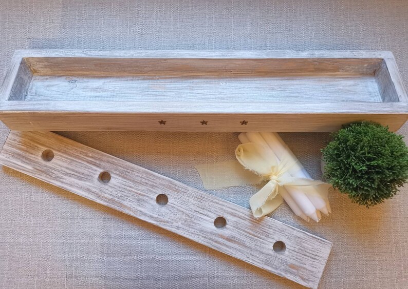 Op de afbeelding: Een witgekalkte houten doos met een rechthoekige opening en drie sterren die in de voorkant zijn gegraveerd. De doos staat op een lichtbruin stofoppervlak. Naast de doos staat een houten dienblad met vijf gaten. Een bundel witte kaarsen en een klein groen struikje liggen ook op het oppervlak.