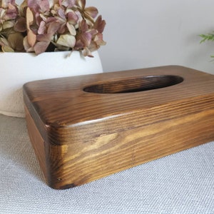 May include: A dark brown wooden tissue box cover with a rectangular shape and a rounded opening for tissues. The wood grain is visible and the cover has a natural, rustic look.