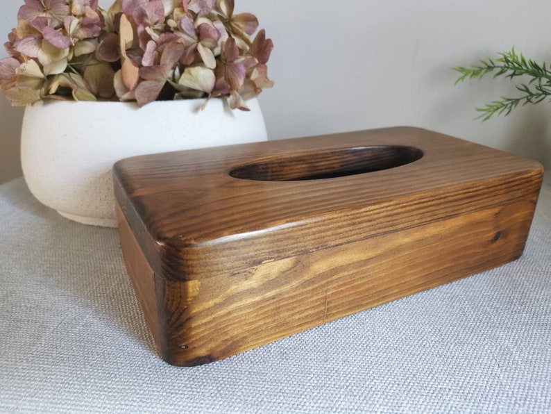 May include: A dark brown wooden tissue box cover with a rectangular shape and a rounded opening for tissues. The wood grain is visible and the cover has a natural, rustic look.