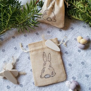 May include: Two small burlap bags with a bunny rabbit design. The bags are filled with small chocolate eggs. The bags are on a white background with green pine branches.