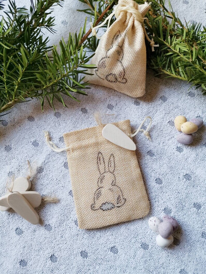 May include: Two small burlap bags with a bunny rabbit design. The bags are filled with small chocolate eggs. The bags are on a white background with green pine branches.