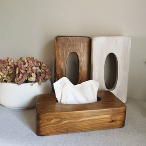 May include: Three wooden tissue box holders. Two are upright and one is laying down with a white tissue inside. The upright holders are stained in a dark brown color and the laying down holder is stained in a lighter brown color.