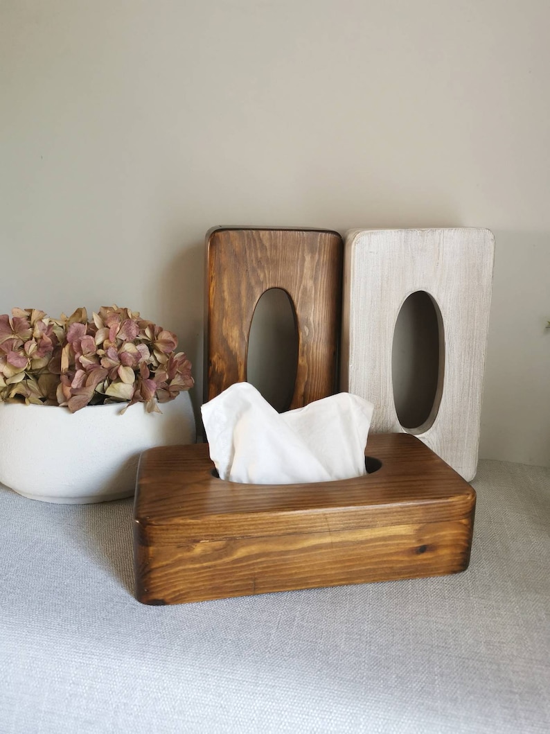 May include: Three wooden tissue box holders. Two are upright and one is laying down with a white tissue inside. The upright holders are stained in a dark brown color and the laying down holder is stained in a lighter brown color.