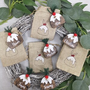 May include: Six brown burlap gift bags with Christmas pudding designs and wooden tags with names: Alex, Brian, Christina, Michael, Jason, and Jeremy.
