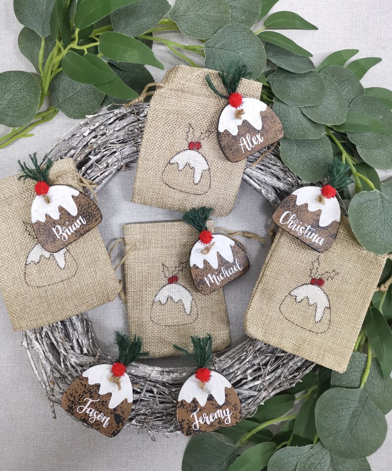 May include: Six brown burlap gift bags with Christmas pudding designs and wooden tags with names: Alex, Brian, Christina, Michael, Jason, and Jeremy.