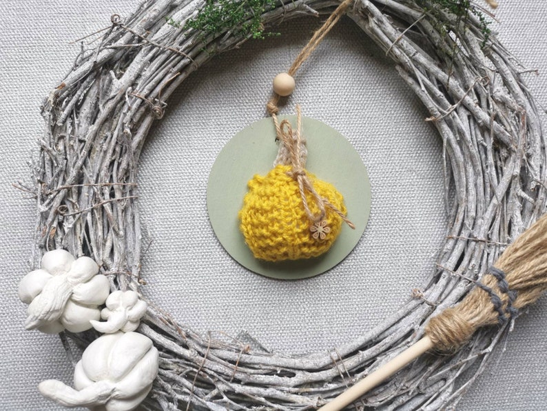 Pu&ograve; includere: Un ornamento a forma di zucca in crochet giallo appeso a una ghirlanda di rami con una piccola perla di legno e uno spago. La ghirlanda &egrave; fatta di rami bianchi e ha una scopa di iuta appoggiata ad essa.