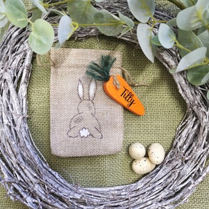 May include: A burlap gift bag with a bunny rabbit design and a carrot-shaped tag that says "Tilly". The bag is surrounded by a twig wreath and speckled Easter eggs.