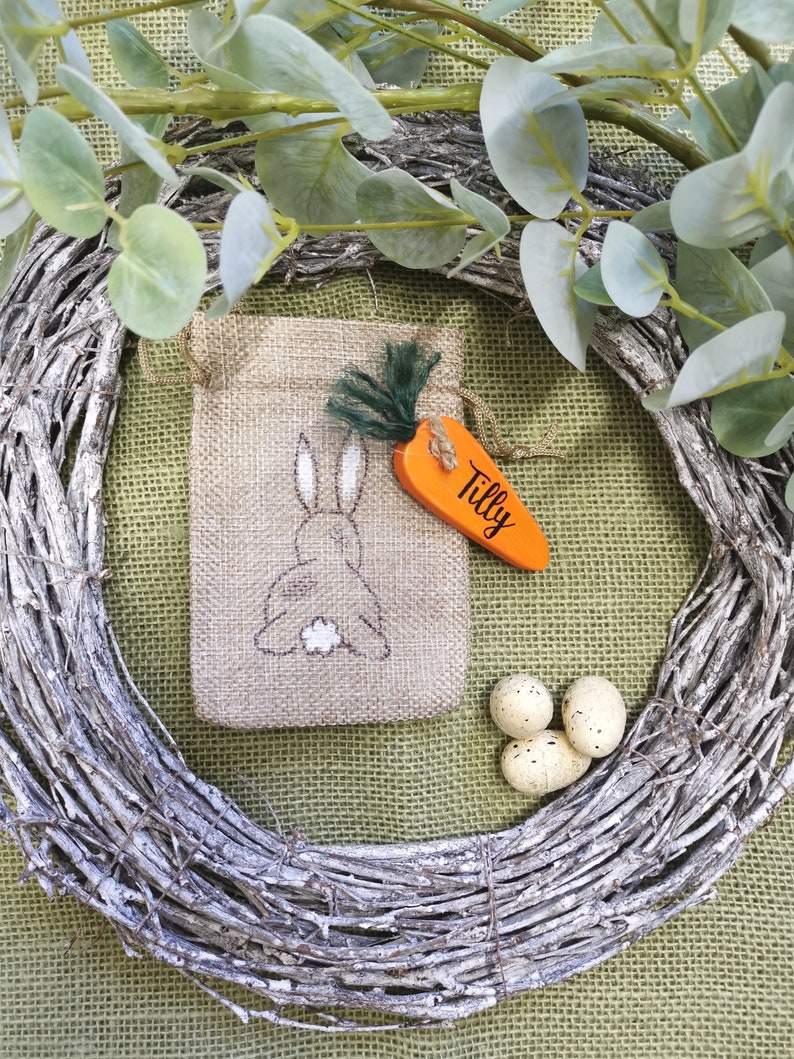 May include: A burlap gift bag with a bunny rabbit design and a carrot-shaped tag that says "Tilly". The bag is surrounded by a twig wreath and speckled Easter eggs.