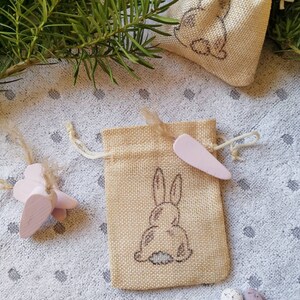 May include: Two small burlap bags with a bunny rabbit design. The bags are filled with small pink and white Easter eggs.