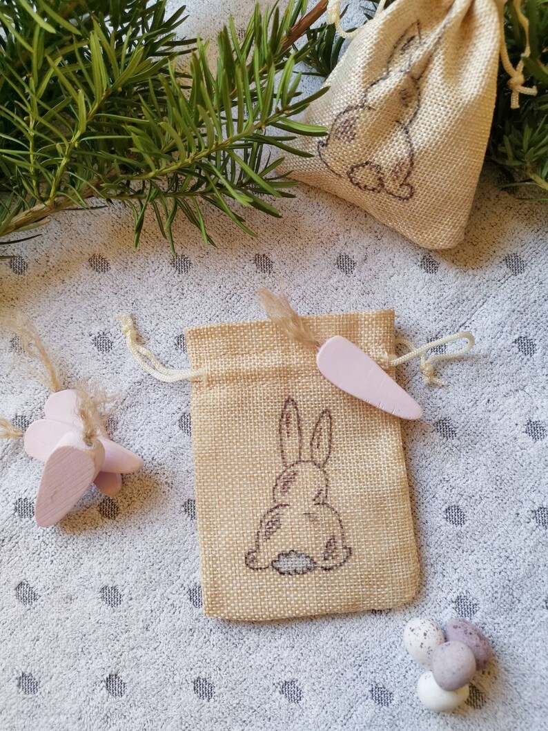 May include: Two small burlap bags with a bunny rabbit design. The bags are filled with small pink and white Easter eggs.