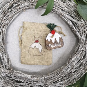 May include: A brown wooden Christmas ornament shaped like a Christmas pudding with white icing and a red candy on top. The ornament is personalized with the name "Michael". The ornament is hanging from a burlap bag with a white drawstring.
