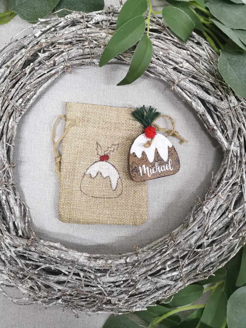 May include: A brown wooden Christmas ornament shaped like a Christmas pudding with white icing and a red candy on top. The ornament is personalized with the name "Michael". The ornament is hanging from a burlap bag with a white drawstring.