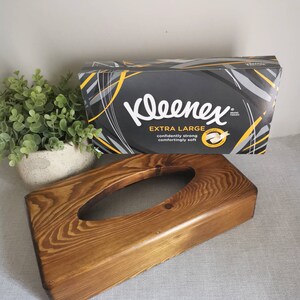 May include: A wooden tissue box holder with a dark brown stain. The holder has a rectangular shape with a rounded top and a cutout in the center for tissues. The box is sitting on a light gray surface.