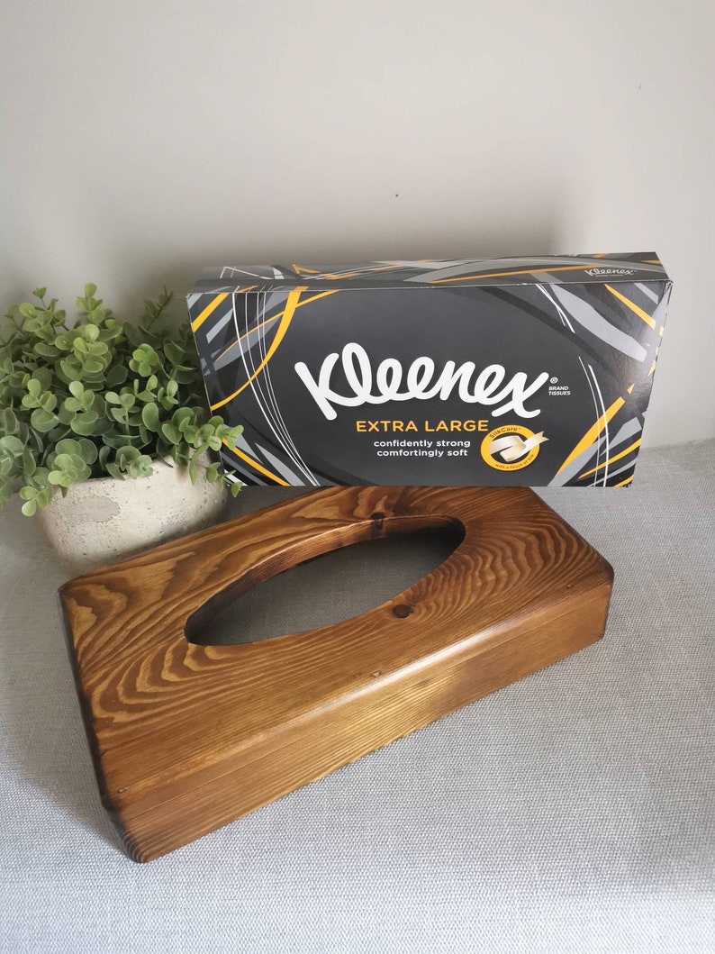 May include: A wooden tissue box holder with a dark brown stain. The holder has a rectangular shape with a rounded top and a cutout in the center for tissues. The box is sitting on a light gray surface.