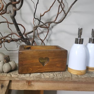 Op de afbeelding: Twee witte keramische zeepdispensers met bamboe bases en zilveren pompen, naast een houten doos met een hartvormige uitsparing. De doos en de dispensers staan op een rustiek houten plank.