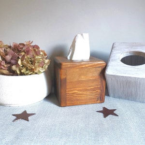 May include: Two tissue box covers, one brown wood and one white painted wood, with a white tissue box inside the brown cover. A white ceramic bowl with dried flowers sits in the background.