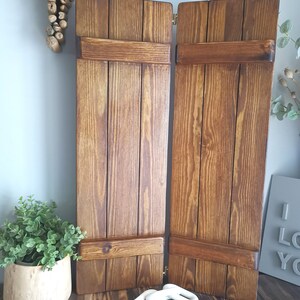 May include: Two wooden shutters with a dark brown stain, each with three horizontal panels and a decorative metal hinge. The shutters are closed and leaning against a wooden surface.