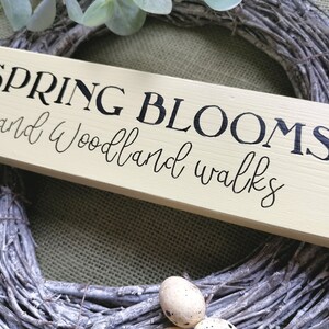 May include: A light yellow wooden sign with black lettering that reads "SPRING BLOOMS and Woodland walks". The sign is sitting on a woven wreath.
