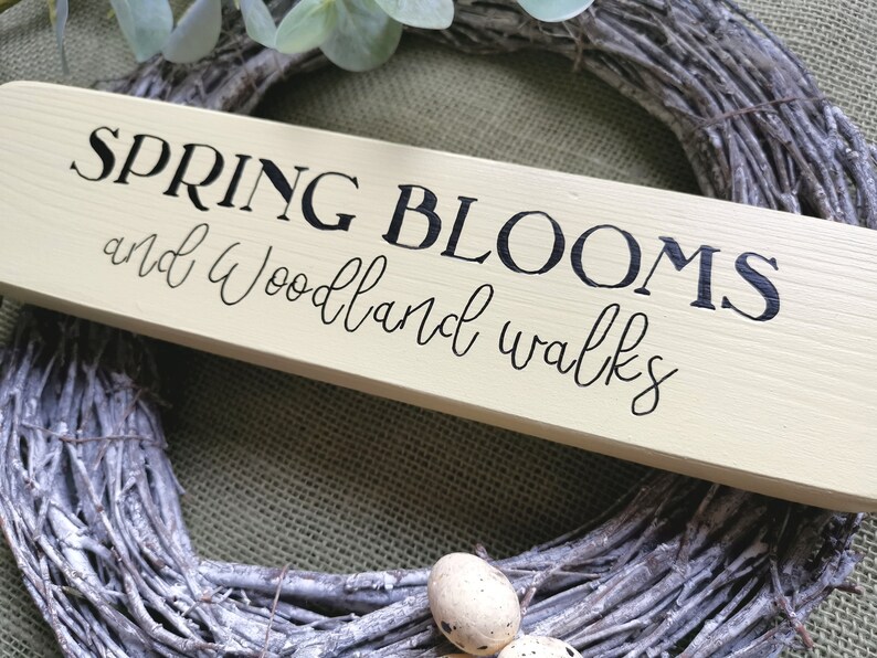 May include: A light yellow wooden sign with black lettering that reads "SPRING BLOOMS and Woodland walks". The sign is sitting on a woven wreath.