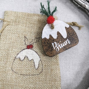 May include: A brown wooden Christmas ornament shaped like a Christmas pudding with white icing and a red candy on top. The ornament has the name "Brian" written on it. The ornament is sitting on a burlap bag with a smaller Christmas pudding design.