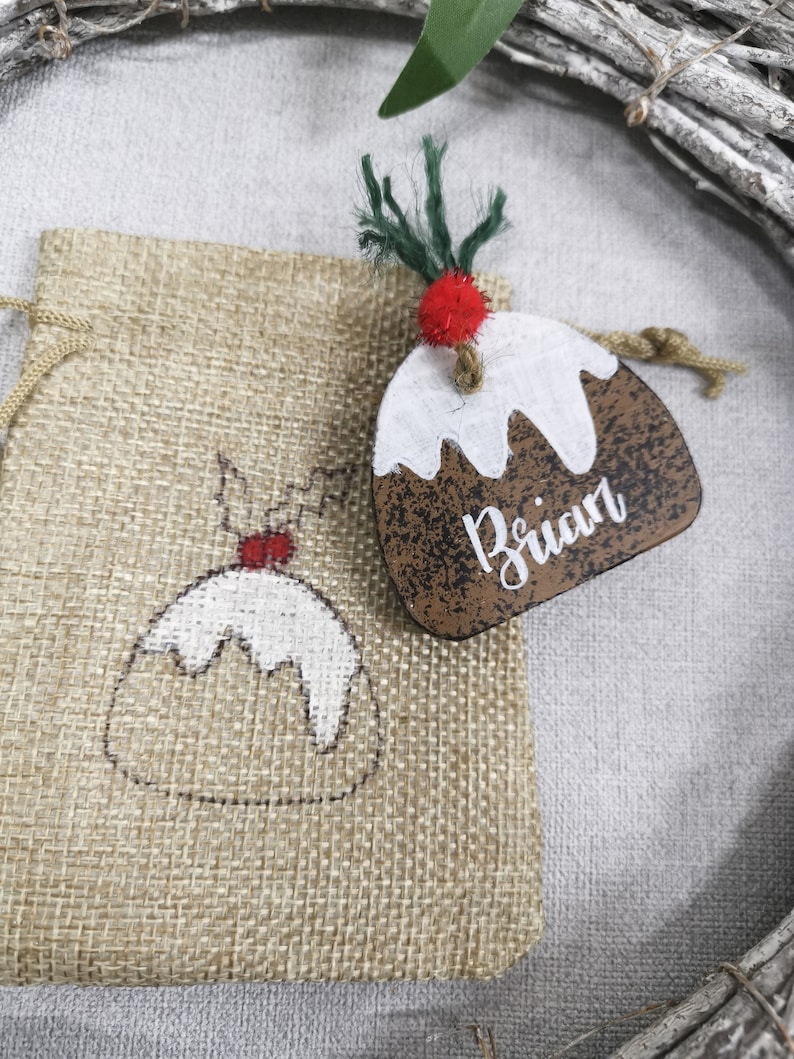 May include: A brown wooden Christmas ornament shaped like a Christmas pudding with white icing and a red candy on top. The ornament has the name "Brian" written on it. The ornament is sitting on a burlap bag with a smaller Christmas pudding design.