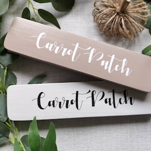 May include: Two wooden signs with the text "Carrot Patch" written in cursive. The top sign is light brown with white lettering. The bottom sign is white with black lettering.
