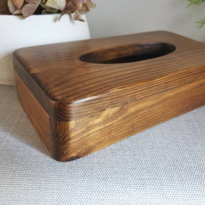 May include: A wooden tissue box holder with a dark brown stain. The box has a rectangular shape with rounded edges and a cutout for tissues.