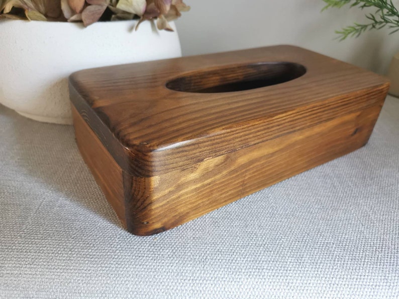 May include: A wooden tissue box holder with a dark brown stain. The box has a rectangular shape with rounded edges and a cutout for tissues.