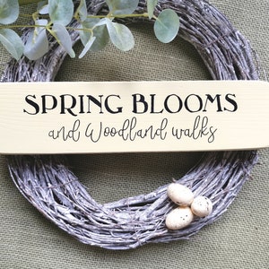May include: A wooden sign with the text "SPRING BLOOMS and woodland walks" in black lettering on a light beige background. The sign is sitting on a burlap surface with a white twig wreath and white speckled eggs.
