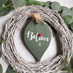 May include: A white twig wreath with a green wooden leaf-shaped ornament that says "Believe" in white lettering with a red star and a red pom-pom.
