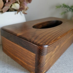 May include: A dark brown wooden tissue box cover with a rectangular shape and a rounded top. The cover has a rectangular opening in the top for tissues.