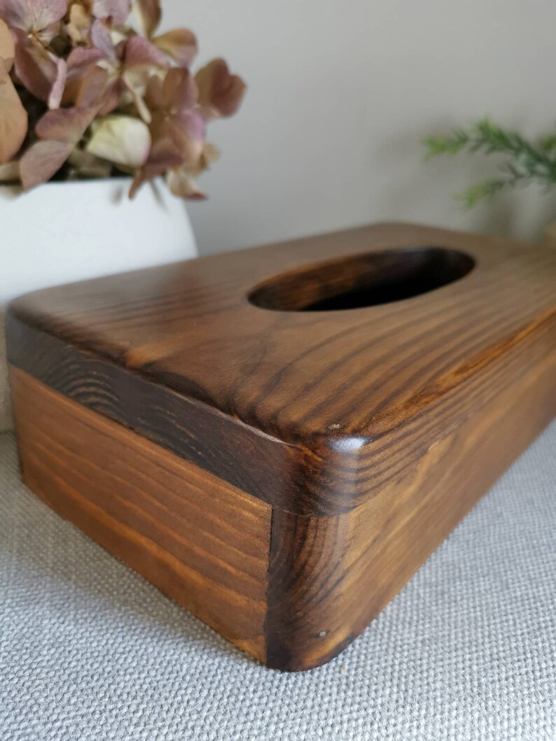 May include: A dark brown wooden tissue box cover with a rectangular shape and a rounded top. The cover has a rectangular opening in the top for tissues.