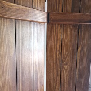 May include: Two wooden shutters, each with four vertical panels, are closed and hinged together. The shutters are a dark brown color with a natural wood grain pattern.